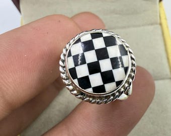 Könnte beinhalten: Ein silberner Ring mit einem runden, karierten schwarz-weißen Design. Der Ring hat einen gedrehten, seilartigen Rand um das Karomuster. Der Ring wird von einer Hand gehalten.