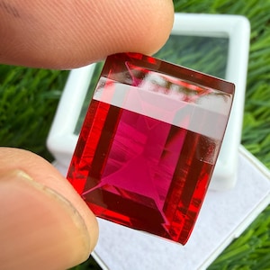 May include: A rectangular, faceted gemstone with a vibrant, deep red color. The stone features a clear, polished surface with sharp, geometric edges and a symmetrical cut that reflects light, showcasing its transparent and saturated red hue.