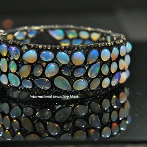 May include: A silver bangle bracelet with a row of opal gemstones set in a black metal frame. The bracelet is reflected in a black surface.