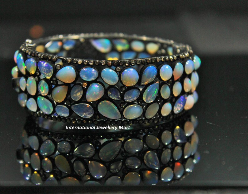 May include: A silver bangle bracelet with a row of opal gemstones set in a black metal frame. The bracelet is reflected in a black surface.
