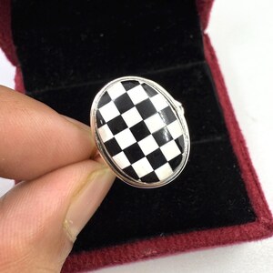 May include: Oval-shaped ring with a black and white checkered design. The ring has a silver-colored band and a checkerboard pattern on the face. The ring is set against a dark red velvet background.