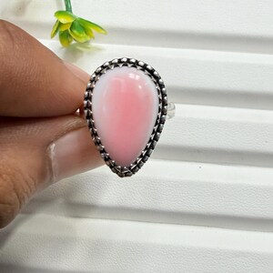 May include: A teardrop-shaped pink stone ring set in a decorative silver metal band with a beaded edge. The smooth, light pink cabochon is securely held within the textured silver setting, showcasing a polished finish and a classic teardrop silhouette.