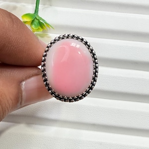 May include: A ring featuring a large, smooth, oval-shaped pink and white stone set in a decorative silver metal bezel with a beaded edge. The stone displays a soft gradient of colour, transitioning from a pale white border to a vibrant pink centre.