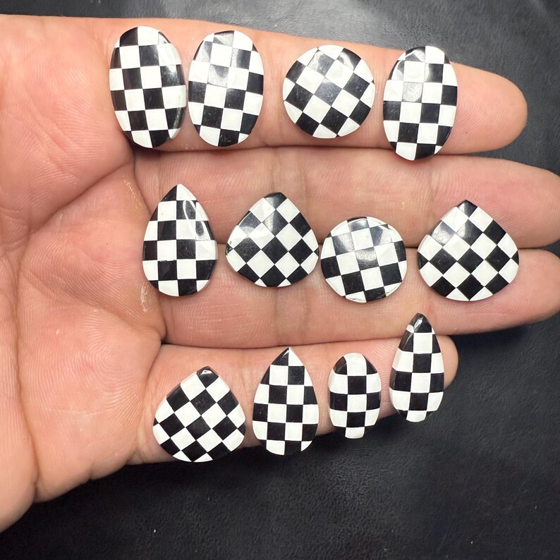 May include: A collection of black and white checkered nail art decorations in various shapes, including ovals, circles, and teardrops. The pieces are arranged on a hand, showcasing the different designs.
