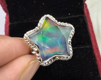Puede incluir: Un anillo en forma de estrella con una gran piedra preciosa iridiscente. La piedra preciosa muestra un espectro de colores, incluyendo verde, azul y púrpura. El anillo tiene un borde plateado retorcido en forma de cuerda. Sobre un fondo de terciopelo rojo oscuro.