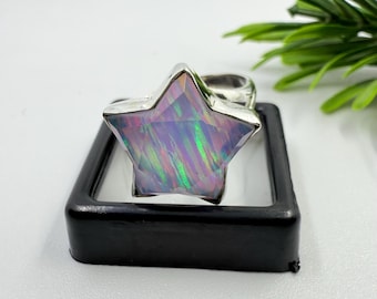 Puede incluir: Anillo de plata con una gran gema en forma de estrella, con colores iridiscentes de púrpura, verde y rosa. El anillo se presenta en una caja cuadrada negra, destacando su diseño único y artesanía.