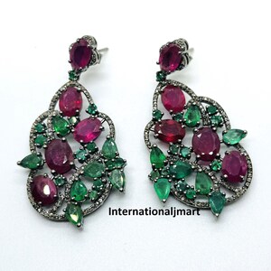 Puede incluir: Un par de pendientes colgantes con un diseño de filigrana plateada. Los pendientes están adornados con rubíes rojos y esmeraldas verdes. Los pendientes están acentuados con pequeños diamantes.