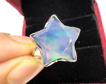 Puede incluir: Un anillo en forma de estrella con una piedra preciosa facetada e iridiscente. La piedra preciosa muestra una gama de colores, incluyendo azul, verde y morado. La banda del anillo es de color plateado. El anillo se muestra sobre un fondo oscuro.