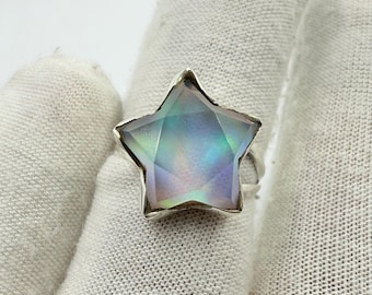Puede incluir: Un anillo de plata con una gran piedra preciosa iridiscente en forma de estrella. La piedra preciosa muestra una gama de colores, incluyendo azules, verdes y morados. El anillo está engarzado en una sencilla banda de plata.