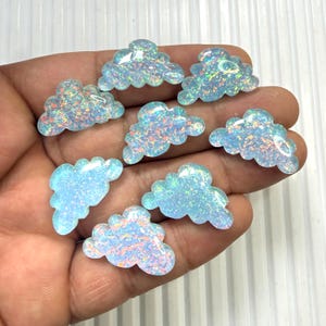 May include: Eight iridescent cloud-shaped cabochons in varying sizes. The cabochons are a light blue color with flecks of rainbow colors throughout. The cabochons are held in a hand against a white background. These are suitable for jewelry making or other craft projects.