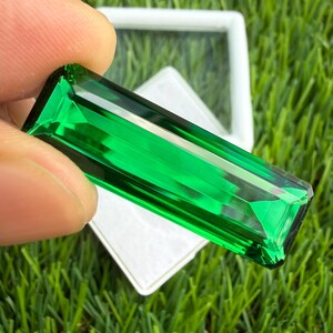 May include: A rectangular, faceted gemstone with a vibrant, translucent emerald green color. The stone features sharp, clean edges and a polished surface that reflects light, showcasing a clear, deep green hue throughout its elongated shape.