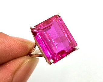 以下が含まれることがあります： 鮮やかなピンク色の宝石リング。長方形にカットされた大きなファセットストーンが、シルバーカラーのバンドにセットされています。白い背景にリングが配置され、宝石の鮮やかな色と透明度を際立たせています。