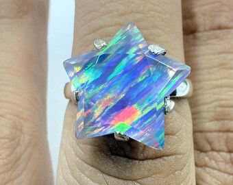 Puede incluir: Anillo de plata con una gran gema en forma de estrella con tonos iridiscentes azules, verdes y naranjas. La gema está facetada, capturando la luz y mostrando un espectro de colores. La banda del anillo es de plata pulida.