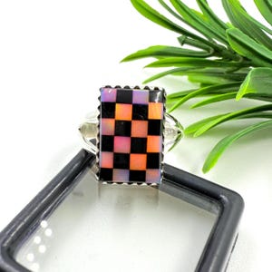 May include: A silver ring with a rectangular, chequered stone. The pattern features black, orange, pink, and purple squares. The ring has a decorative, scalloped edge around the stone.