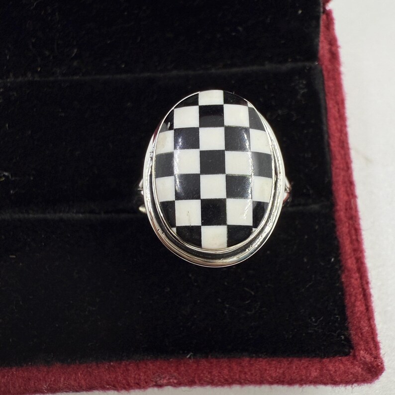 May include: Oval-shaped ring with a black and white checkered design. The ring has a silver-colored band and is set against a black velvet background.