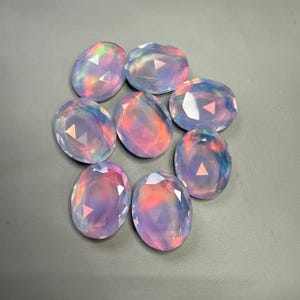 May include: Eight oval-shaped gemstones with a faceted cut. The gems display a range of colors, including pink, purple, and blue, with iridescent flashes. The stones are arranged on a gray surface, showcasing their polished appearance.
