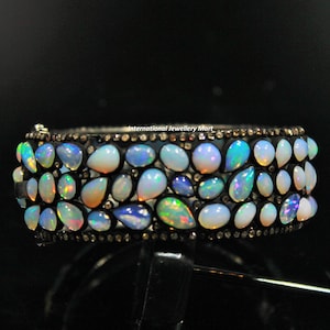 May include: A silver bangle bracelet with a row of iridescent opal gemstones set in a black metal band. The bracelet is adorned with small black diamonds.