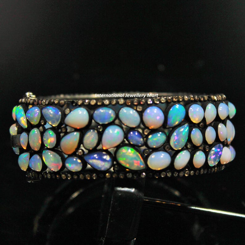 May include: A silver bangle bracelet with a row of iridescent opal gemstones set in a black metal band. The bracelet is adorned with small black diamonds.