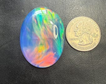 May include: An oval-shaped opal gemstone with a vibrant display of colors, including blue, green, orange, and pink. The gemstone is next to a U.S. quarter for size comparison. The opal has a polished, glossy surface.