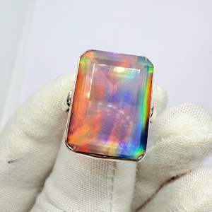Può includere: Un anello d'argento con una grande pietra rettangolare e iridescente. La pietra preziosa presenta un arcobaleno di colori, tra cui rosa, arancio, giallo, verde e blu.