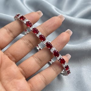 Puede incluir: Una pulsera de tenis en tono plateado que presenta un patrón repetitivo de piedras de color rojo intenso con corte ovalado, alternadas con pequeñas piedras de corte redondo y transparentes. La pulsera tiene un diseño de eslabones metálicos flexibles.