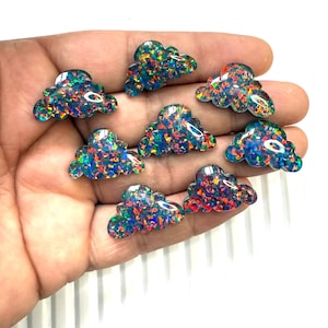 May include: A collection of eight cloud-shaped hair clips. Each clip is a translucent, iridescent material with a mix of blue, green, red, and yellow flecks. The clips are held in a hand against a white background.