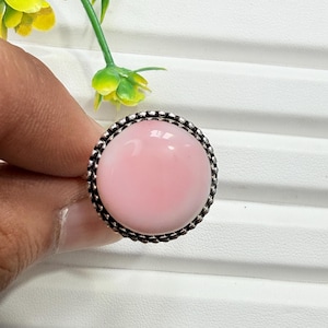 Puede incluir: Un anillo con una piedra cabujón grande, lisa, redonda y de color rosa claro, engastada en una banda de metal de tono plateado oscuro y oxidado con un borde texturizado y con cuentas que rodea la gema, creando un contraste entre el rosa y el plata.
