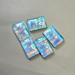 Puede incluir: Seis ópalos rectangulares con un brillo iridiscente azul, verde y naranja. Los ópalos están dispuestos sobre una superficie gris. Las piedras tienen un aspecto pulido y vidrioso, y la luz se refleja en sus superficies.