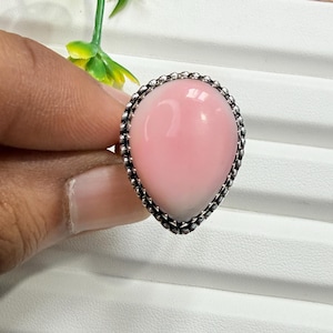 May include: A teardrop-shaped pink stone ring set in a silver metal band with a beaded edge. The smooth, polished pink gemstone is centred within the decorative silver setting, which features a textured, dotted border around the perimeter.