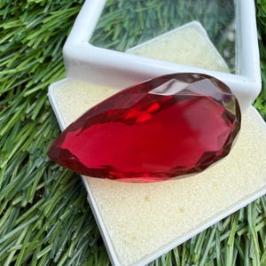 Può includere: Una gemma rossa sfaccettata a forma di pera, appoggiata su un inserto in schiuma bianca all'interno di una scatola espositiva in plastica bianca. La pietra rosso intenso presenta molteplici sfaccettature geometriche.
