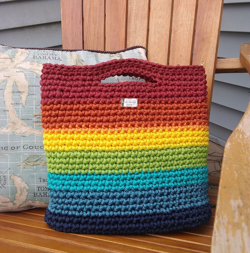 Pu&ograve; includere: Una borsa a mano all'uncinetto con un motivo arcobaleno. La borsa &egrave; realizzata con una variet&agrave; di colori, tra cui rosso, arancione, giallo, verde, blu e viola. La borsa ha un'unica maniglia e un piccolo cartellino che dice "Fatto a mano".
