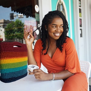 Pu&ograve; includere: Una persona con un vestito arancione brillante &egrave; seduta a un tavolo bianco con una borsa all'uncinetto arcobaleno e una coppetta di gelato. La persona sorride e guarda a destra.