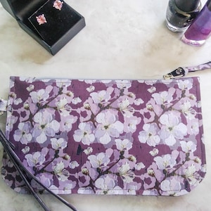 May include: A purple floral clutch with a wrist strap, featuring a white flower pattern. The clutch is rectangular and has a zipper closure. A pair of pink gemstone stud earrings in a black box and two bottles of nail polish are also visible.
