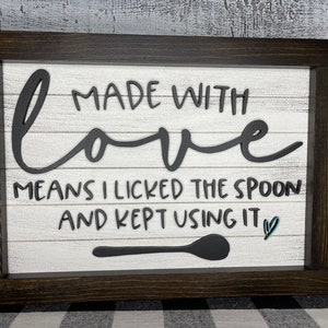 May include: A white wooden sign with black lettering that reads "Made with love means I licked the spoon and kept using it". A black wooden spoon is pictured below the text with a small blue heart.