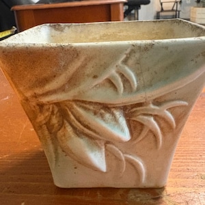 May include: A square-shaped ceramic planter with a tapered base, featuring a raised relief design of leaves and stems. The surface has a cream-coloured glaze with brown, weathered-looking accents and visible crackling throughout the exterior.