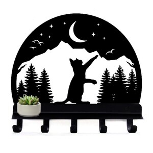 Puede incluir: Porta llaves de metal negro con una silueta de un gato alcanzando una luna creciente en un paisaje de montaña. El porta llaves tiene cinco ganchos y una pequeña repisa para una planta en maceta.