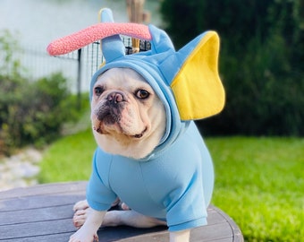 May include: A white French bulldog wearing a blue and yellow elephant costume. The dog is sitting on a brown wooden surface.