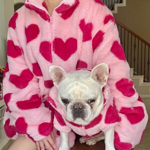 Puede incluir: Una chaqueta de forro polar rosa y roja con estampado de corazones. Un bulldog franc&eacute;s blanco lleva una chaqueta a juego.