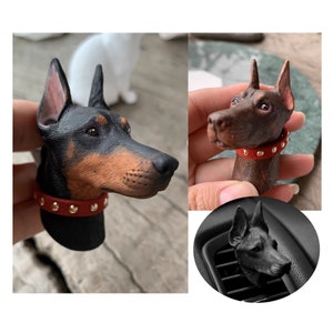Könnte beinhalten: Zwei schwarze und braune Dobermann-Hunde-Kopf-Auto-Lufterfrischer-Ornamente mit roten Halsbändern und goldenen Nieten. Einer wird in der Hand gehalten, der andere befindet sich in einer Lüftung des Autos.