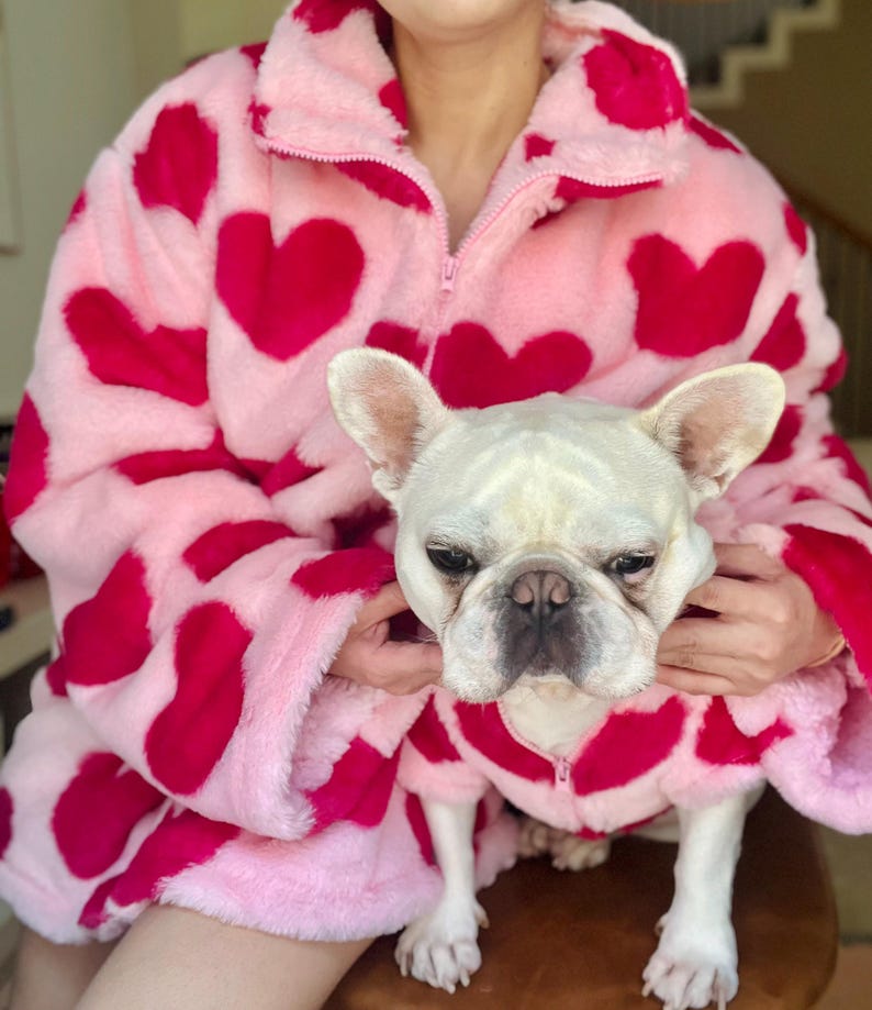 Puede incluir: Una chaqueta de forro polar rosa y roja con estampado de corazones. Un bulldog franc&eacute;s blanco lleva una chaqueta a juego.
