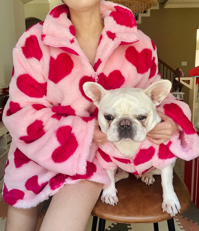 Puede incluir: Una chaqueta de forro polar rosa y roja con estampado de corazones y una chaqueta para perro a juego. La chaqueta la lleva una persona y la chaqueta para perro la lleva un peque&ntilde;o bulldog franc&eacute;s blanco.