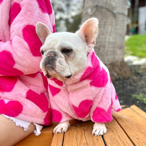 Puede incluir: Un bulldog franc&eacute;s blanco con una chaqueta de forro polar rosa con estampado de corazones. La chaqueta tiene una cremallera y est&aacute; hecha de tela suave y afelpada.