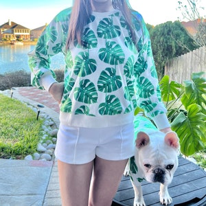 May include: A white sweatshirt with a green tropical leaf print. The sweatshirt is worn by a person with white shorts and a white dog wearing a matching green tropical leaf print shirt. The dog is a French Bulldog.
