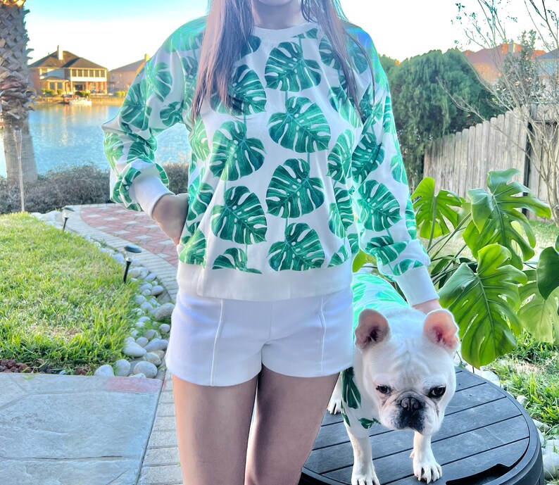 May include: A white sweatshirt with a green tropical leaf print. The sweatshirt is worn by a person with white shorts and a white dog wearing a matching green tropical leaf print shirt. The dog is a French Bulldog.