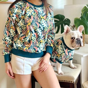 K&ouml;nnte beinhalten: Eine Frau und ein Hund tragen passende Sweatshirts mit tropischem Muster in Gr&uuml;n und Gelb. Die Frau tr&auml;gt wei&szlig;e Shorts und der Hund sitzt auf einem Hocker.