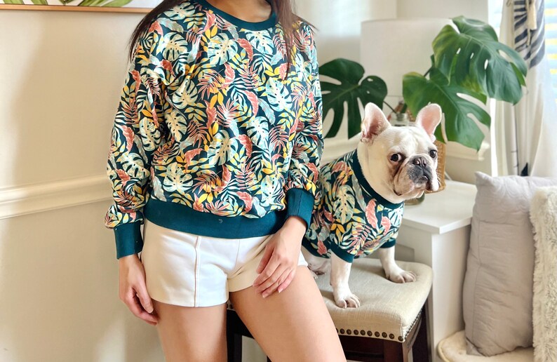 K&ouml;nnte beinhalten: Eine Frau und ein Hund tragen passende Sweatshirts mit tropischem Muster in Gr&uuml;n und Gelb. Die Frau tr&auml;gt wei&szlig;e Shorts und der Hund sitzt auf einem Hocker.