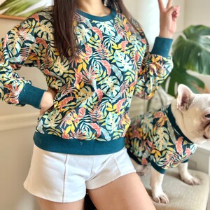 K&ouml;nnte beinhalten: Ein blaugr&uuml;ner Pullover mit tropischem Bl&auml;tterdruck. Der Pullover hat einen Rundhalsausschnitt und lange &Auml;rmel. Ein wei&szlig;er Hund, der ein passendes Shirt tr&auml;gt, steht neben der Person, die den Pullover tr&auml;gt.