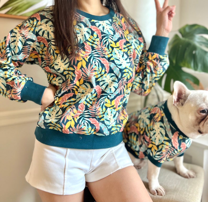 K&ouml;nnte beinhalten: Ein blaugr&uuml;ner Pullover mit tropischem Bl&auml;tterdruck. Der Pullover hat einen Rundhalsausschnitt und lange &Auml;rmel. Ein wei&szlig;er Hund, der ein passendes Shirt tr&auml;gt, steht neben der Person, die den Pullover tr&auml;gt.
