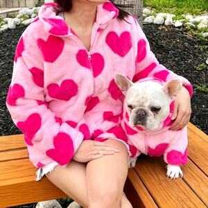 Puede incluir: Una chaqueta de color rosa peluda con corazones rosas impresos en ella. Un bulldog franc&eacute;s blanco lleva una chaqueta de color rosa peluda a juego con corazones rosas impresos en ella.