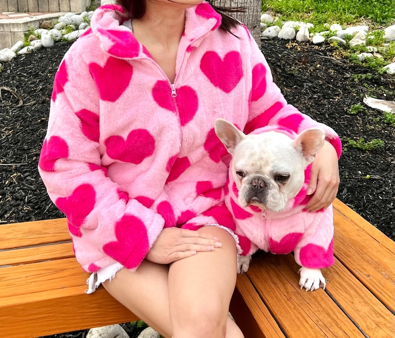 Puede incluir: Una chaqueta de color rosa peluda con corazones rosas impresos en ella. Un bulldog franc&eacute;s blanco lleva una chaqueta de color rosa peluda a juego con corazones rosas impresos en ella.